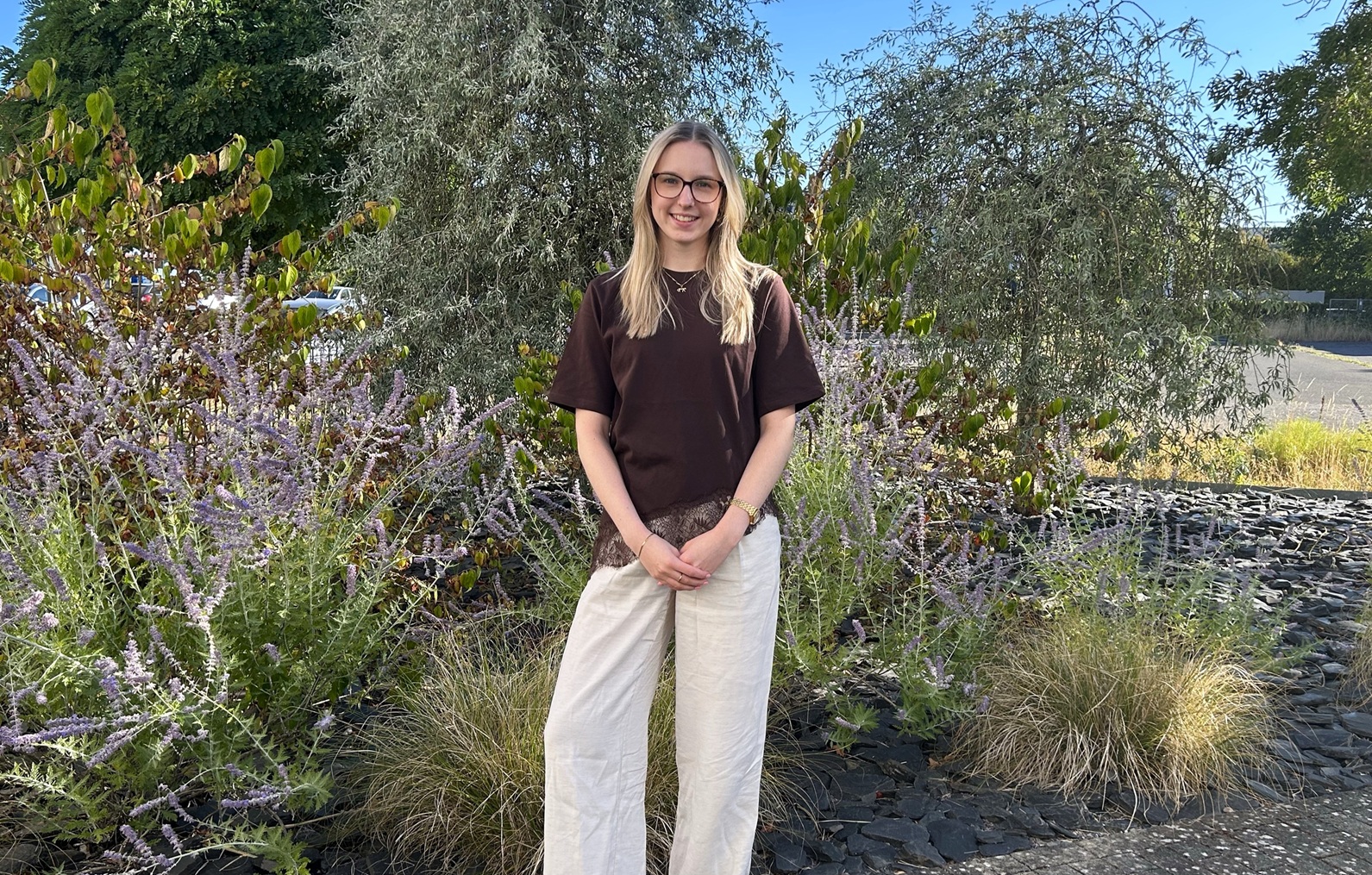 Eine blonde frau steht in einem Garten mit Pflanzen und lächelt in die Kamera.